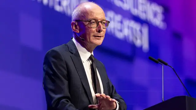 Pat McFadden speaking in front of a microphone, wearing a black suit and tie