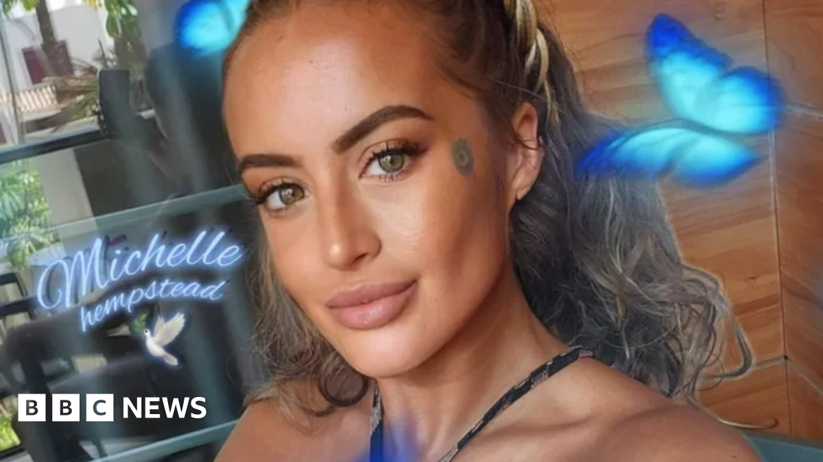 Michelle Hempstead has dark, curly hair swept back behind her head. This picture is a selfie that has cartoon blue butterflies and her name printed on it. Michelle is smiling with her mouth closed in front of wooden panels.