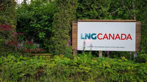 Bloomberg via Getty Images The entrance to the LNG Canada facility in Kitimat, British Columbia, Canada, shows a white sign reading 'LNG Canada' in blue and red and in smaller print 'located in the traditional territory of the Haida Nation'. 
In the background are lush green bushes and trees.