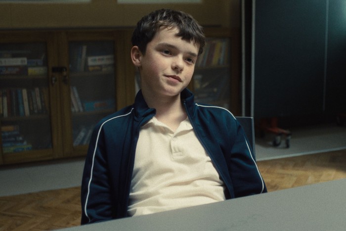 Owen Cooper wearing a white polo shirt and blue jumper, smiling and looking ahead.