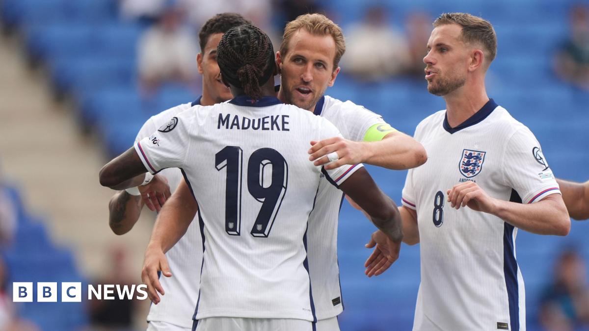 Harry Kane is about to hug his teammate, Noni Madueke. They are wearing white England football shirts with navy blue accents on them. Harry Kane is wearing a bright green captain's armband. Next to Harry is Jordan Henderson, moving in for a hug.