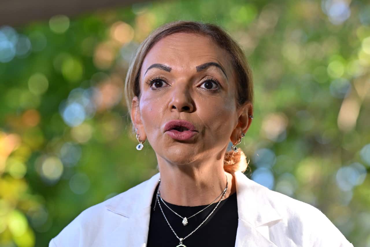 Anne Aly wearing a white shirt speaking in front of green trees.