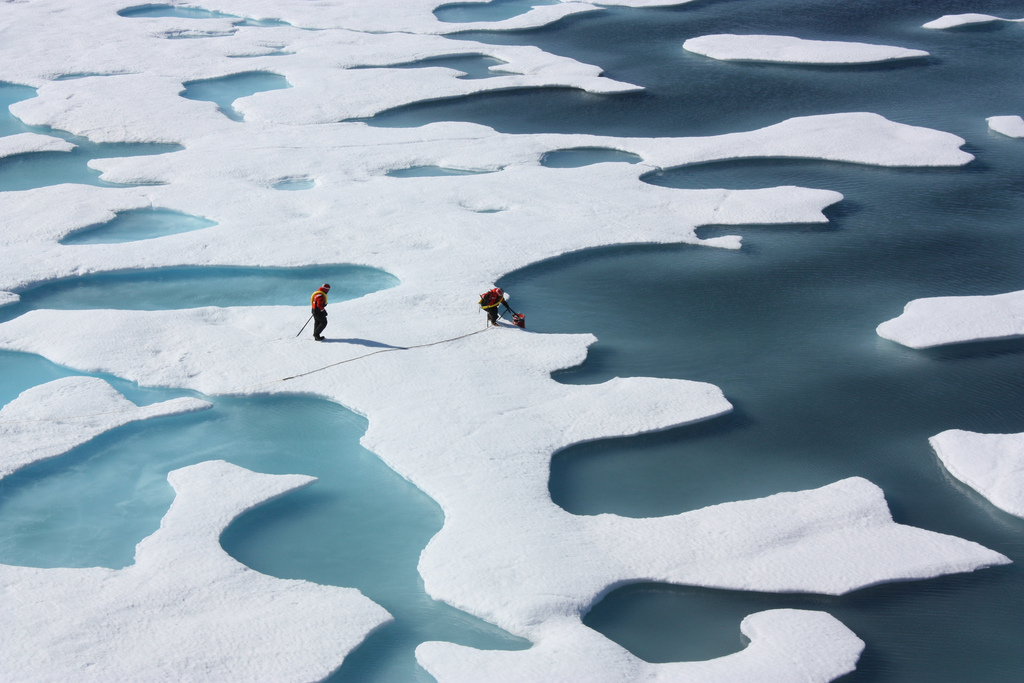 Two researchers in bright jackets walk across fragmented Arctic sea ice, navigating around pools of melting blue water, illustrating the thinning and melting ice in the polar region. Credit Kathryn Hansen - NASA