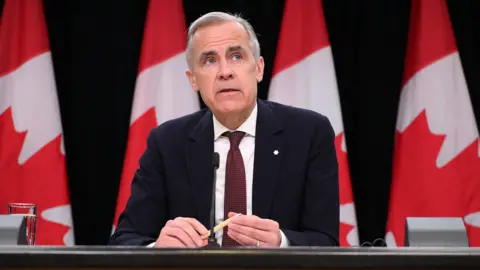 Bloomberg via Getty Images Canada's PM Mark Carney looks to the right of frame while sitting at a press conference briefing table in front of four Canadian flags in Ottowa in May. He wears a black suit and red tie, and holds a pen in his hands with a glass of water beside him on the table.