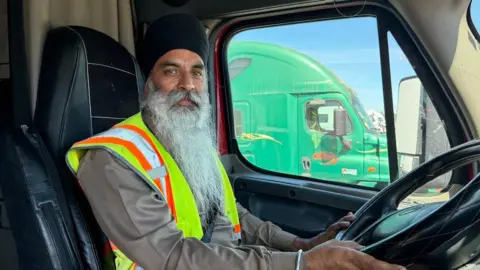 Gagandeep Singh A truck driver, with a long white beard, sits behind the wheel in his truck cab. He is wearing a yellow safety vest and a black turban. Through the window, you can see another green truck.