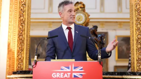 Reuters Business and Trade Secretary Peter Kyle hosts a business reception at Lancaster House in central London. He wears a navy suit with a white shirt and a burgundy tie. He is standing in front of an opulent, gold-framed mirror and in front of a red government podium enblazoned with a Union flag reading Great Britain and Norhern Ireland.
