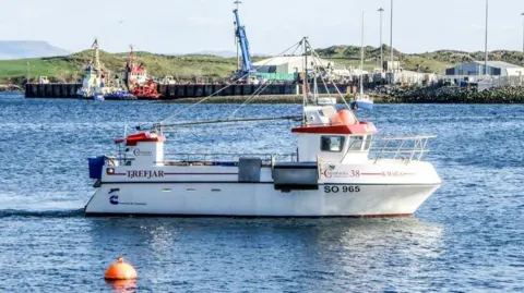The Skipper magazine Kevin McCloskey's fishing boat, the FV K-Mar-K, on the water near a rural harbour on a sunny day.  The boat is white with red branding. There is a red canopy over the wheelhouse and there is a metal safety rail around the hull. 