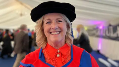 BBC Gillian wears a black graduation cap and red and blue gown. She is smiling at the camera. She is in a marquee, with people behind her out of focus.