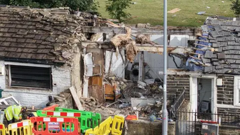 YappApp An close up shot of a terraced house with extensive explosion damage including a decimated roof and debris all over the road and back garden