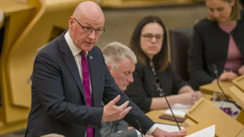 PA Media John Swinney, a bald man with glasses, speaks at a podium in the Scottish Parliament. He is wearing a dark suit, white shirt and purple tie, with his right hand gesticulating in front of his body. There are MSPs sat on benches behind him. 