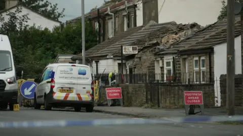 A Northern Gas Network van parked on a quiet street behind blue and white police tape. There are red 'footpath closed' signs on the pavement. A person in a yellow jacket can be seen in the distance working on rubble.