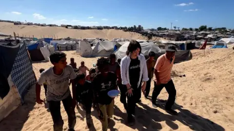 A group of people walks up a slope in al-Mawasi. One is carrying a blue bowl. Behind them are several rows of tents and makeshift dividers. It is daytime.