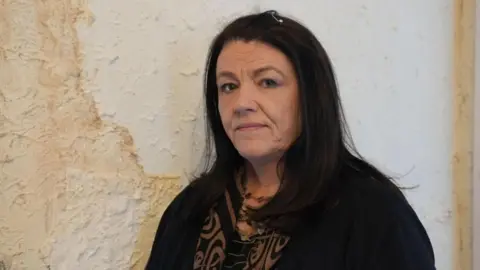 Susan Haslam with long dark hair and a black cardigan and brown and black patterned blouse looks at the camera with a negative expression as she stands in front of a white wall with a huge patch of brown damp