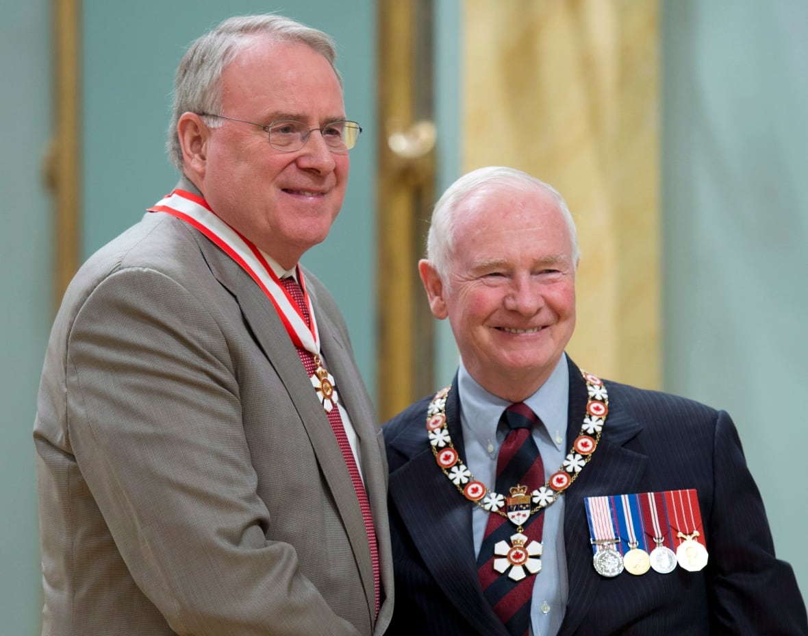 A tall grey-haired man wearing glasses and a grey suit, with a medal hanging around his neck, shakes hands with a white-haired man in a dark suit displaying medals.