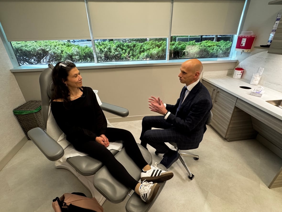 A woman wearing all black sits on a chair with her legs elevated, as a man sitting beside her is seen talking to her.