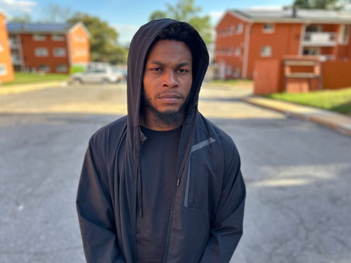 Bryan Turner is a black man with short black hair and a goatee. He is wearing a black nylon sport jacket with the hood over his head. He has on a black tshirt and is standing in the middle of a parking lot. You can see three red brick buildings behind him.