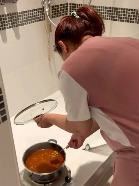 A woman in a pink and white t-shirt cooks over an electric hob on the floor of a shower. She has her dyed red hair tied up. The shower is white with a row of grey tiles. She is holding the lid of the hob in her left hand and stirring a red mixture with the other.