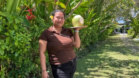 Ade Mardiyati Ni Kadek Sintya in a brown polo shirt, holding a coconut on a plate. There is a yellow frangipani in her hair and she is smiling.