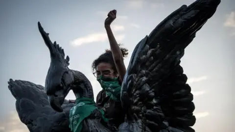 John Reyes Mejia/EPA/Shutterstock A woman who is covering her mouth with a green bandanna lifts her left fist in a gesture of defiance as she is perched on top of a statue during a protest in Lima.
 