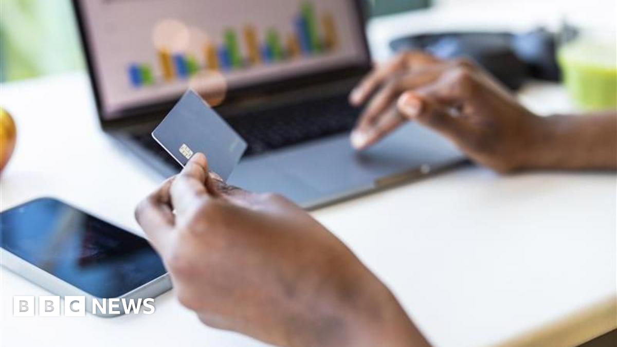 A person's holds a bank card in their left hand, while using a laptop trackpad with their right. On the computer screen are some bar charts.