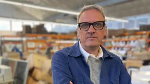 Mark Hargreaves, who runs a trolley and tray manufacturing and export business in Peckham, south London, has his arms folded. He is wearing a blue shirt, a grey jumper and a white shirt. He looks straight faced at the camera and is wearing black-framed glasses.