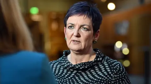 Getty Images Angela Constance, who has short hair that is dyed purple, speaks to a  journalist in the Scottish Parliament. She is photographed over the shoulder of the journalist, and is wearing a black and green patterned top. 