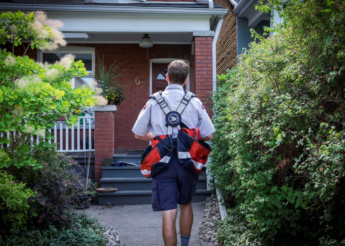 A postal worker is seen from the back, walking toward a brick house