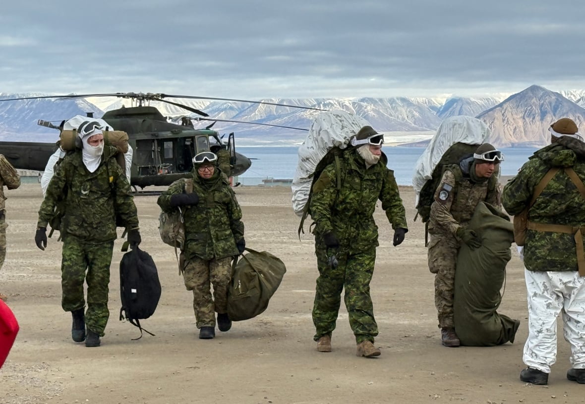 Five soldiers in stand on a barren landscape in heavy winter clothing. There's a helicopter and some mountains in the background.