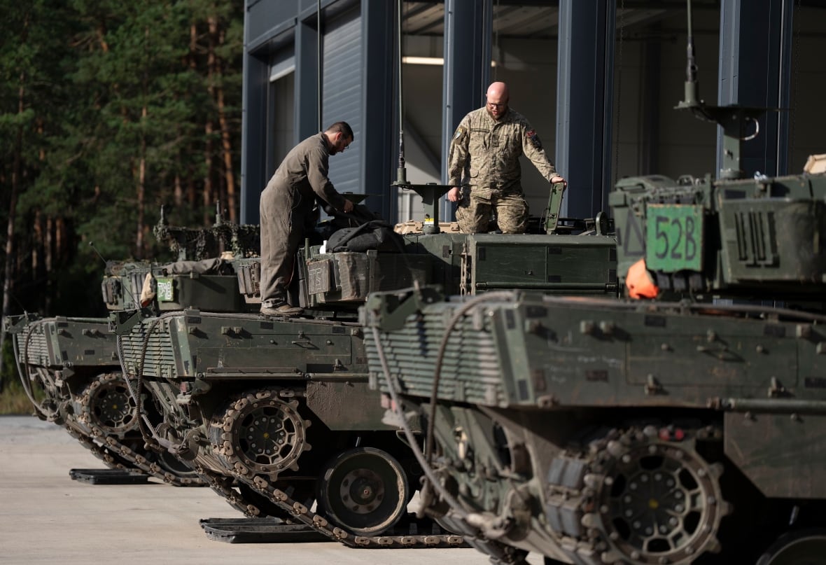 A row of three tanks. Two soldiers in uniform stand on top of the one in the middle.