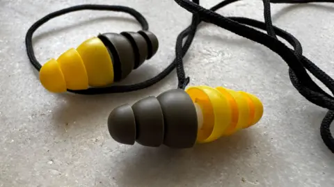 A close up shots of two combat earplugs supplied by a unit of 3M. It shows how they were reversible with yellow and green plastic ends giving different levels of protection. Both earplugs are connected to each other in the photo by a black string worn around the neck.