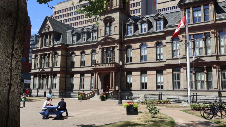 People sit at a blue picnic table on a cobblestone courtyard in front of a three-story historic stone building with a clock tower in the centre There is a Canadian flag on a flagpole to the right and two bikes locked at bike racks. The trunk of a tree is in the foreground to the left.