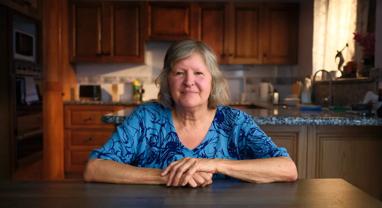 A woman in a blue shirt with her arms folded across a table. She has shoulder length hair and sits in a kitchen.