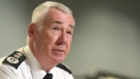 PA Media Jon Boutcher speaks to reporters while wearing his PSNI uniform. He has short white hair and dark eyebrows.
