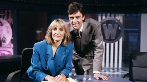 Lynn Faulds Wood in a blue blazer sits behind a desk on the Watchdog set in the 1980s. John Stapleton, in a grey suit, stands behind her. 
