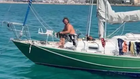 Nikita Goddard James Nunan sitting on the deck of his small green boat. He is topless and smiling while at sea.