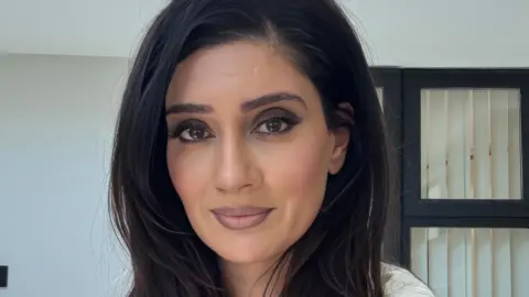 Sophia K Choudry Sophia Choudry smiles in front of a white wall and window whilst wearing a white top