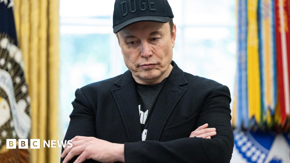 Elon Musk stands in the oval office in a black suit jacket, a black tshirt and a black cap with the letters DOGE stitched in. His arms are folded and he is looking downwards. In the background the flag with the President's standard can be seen to the right, it is out of focus.