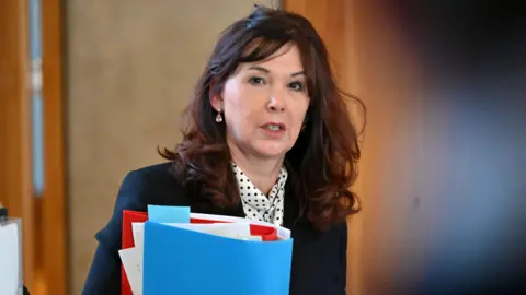Getty Images Dorothy Bain has long brown hair. She is wearing a suit jacket over a white blouse with black dots on it. She is carrying a folder with papers in it.