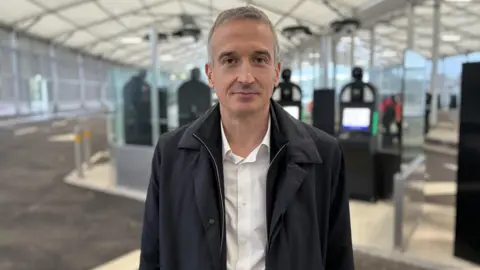 Simon Jones/BBC Close up of Eurotunnel CEO Yann Leriche standing in a hanger in Folkestone with biometric machines behind him. He has short grey hair and is wearing a black jacket and white shirt.
