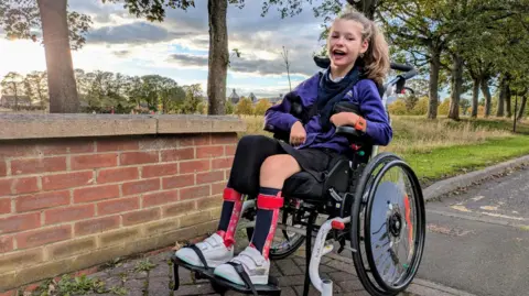 Family handout Wren is looking directly into the camera with a wide smile. She is wearing her school uniform and is sitting in her manual wheelchair. 
Wren's hair is tied up in a ponytail and is wearing leg splints and white trainers.
The picture has been taken in a wooded area.  