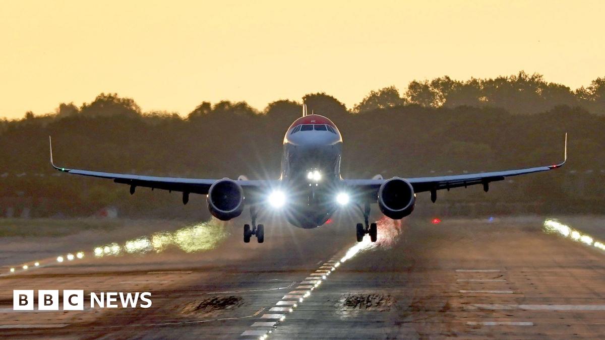 A plane takes off from London Gatwick airport in Crawley, West Sussex - the plane can be seen front a short distance off the ground, either about to land or take off - it is at sunset with the golden glow of the sun lighting up the area around the runway, with silhouetted trees in the background