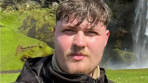 Police Scotland A man with brown hair, a moustache and goatee beard smiles while looking into the camera. He is standing outside while looking into the camera on a sunny day. A waterfall can be seen in the distance behind him.