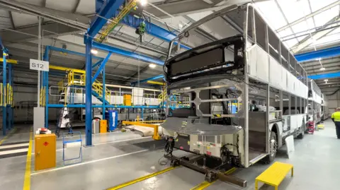 Alexander Dennis The inside of the firm's factory in Larbert. There is the shell of a bus under construction in a large facility with grey walls and blue beams and columns.