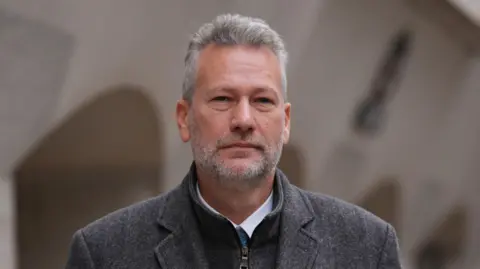 PA Nathan Gill; a man with a grey coat on, and underneath there is a turquoise tie and white shirt. He has short grey spiked hair and a a grey beard.