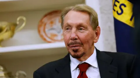 Getty Images Larry's head and shoulders can be seen as he sits in the Oval Office, in front of a shelf with trophies and a decorative plate. He is wearing a black suit jacket, deep red tie, and has a moustache and goatee.