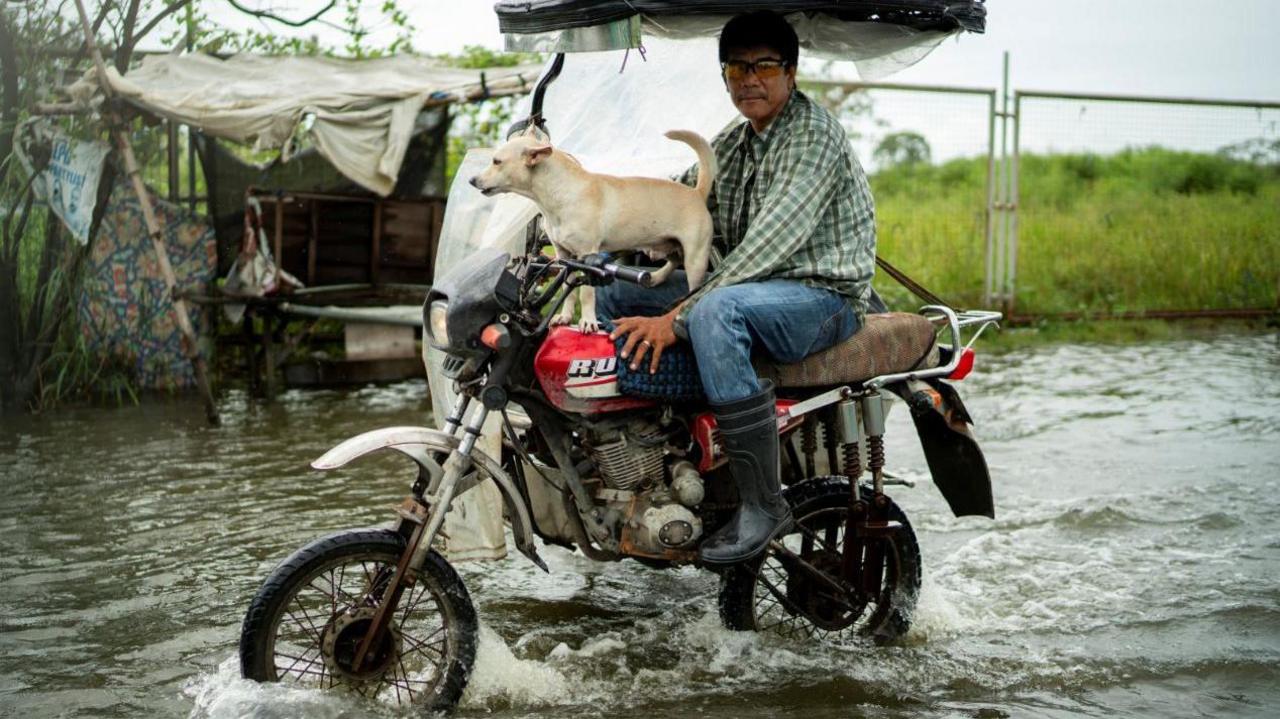 A photo of a man and a dog riding a motorbike through floodwaters