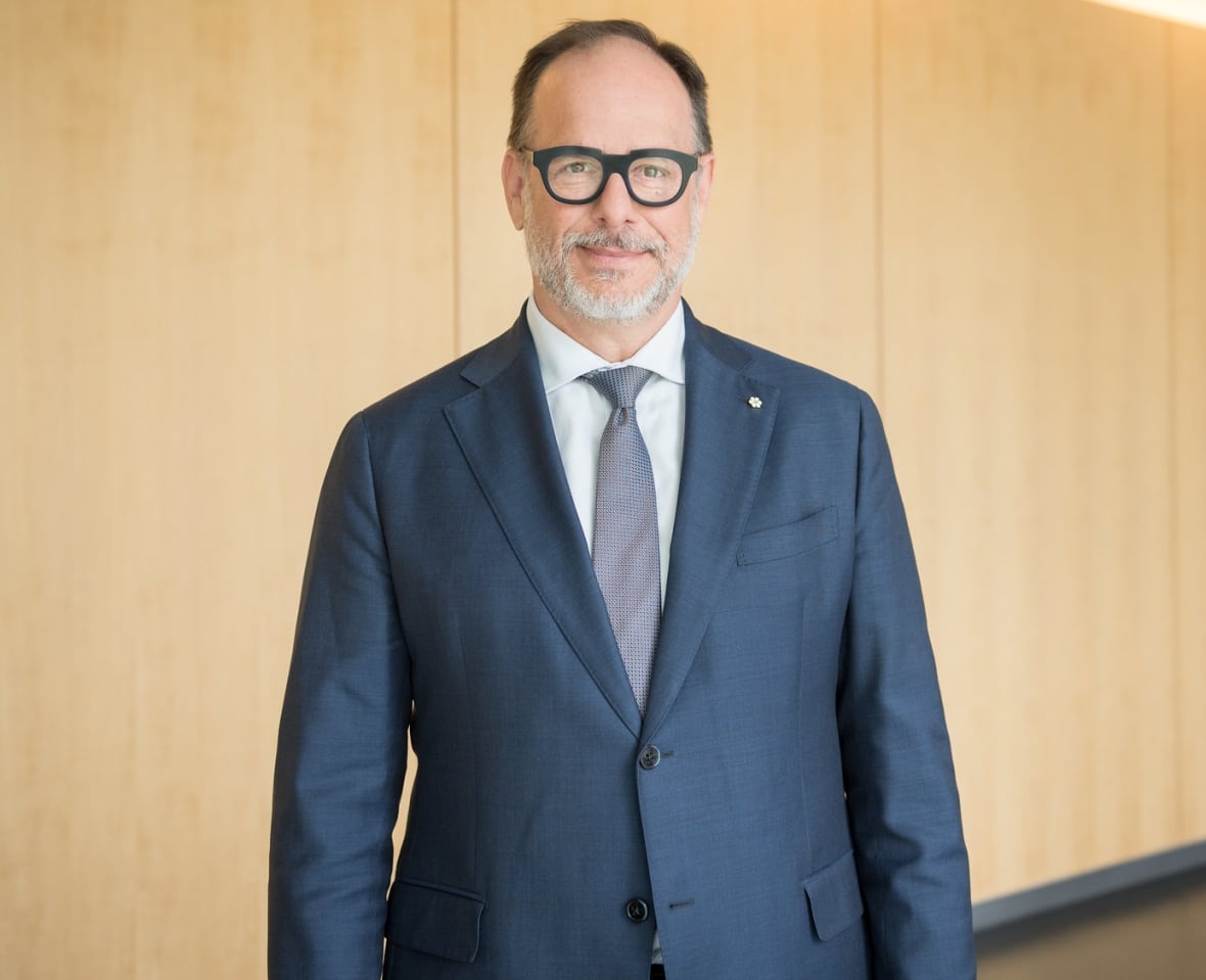 A man in a navy suit, dark-rimmed glasses and a trim beard stands, slightly smiling, in an indoor hallway with wooden panelling. 