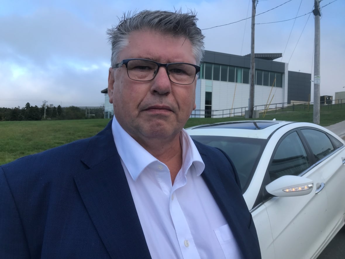 A chest-up closeup of a person wearing glasses, a blazer and a white dress shirt. The person is standing in front of a car and a grey and white building in the distance.