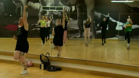 One girl, in a black tank top and black shorts, leads a group of other women in a dance class. She is standing in front of a wall of mirrors. In the reflection behind her, you can see a group of 6 women mimicking her dance moves on a wooden floor. On the wall behind them is a picture of people doing push ups spanning the entire wall. 