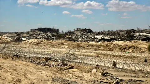 BBC/Dave Bull A coil of barbed wire runs across churned-up piles of soil near the city of Rafah. Demolished buildings lie in piles of debris behind the wire, with one lone structure still upright but appearing to have had its windows blown out.  